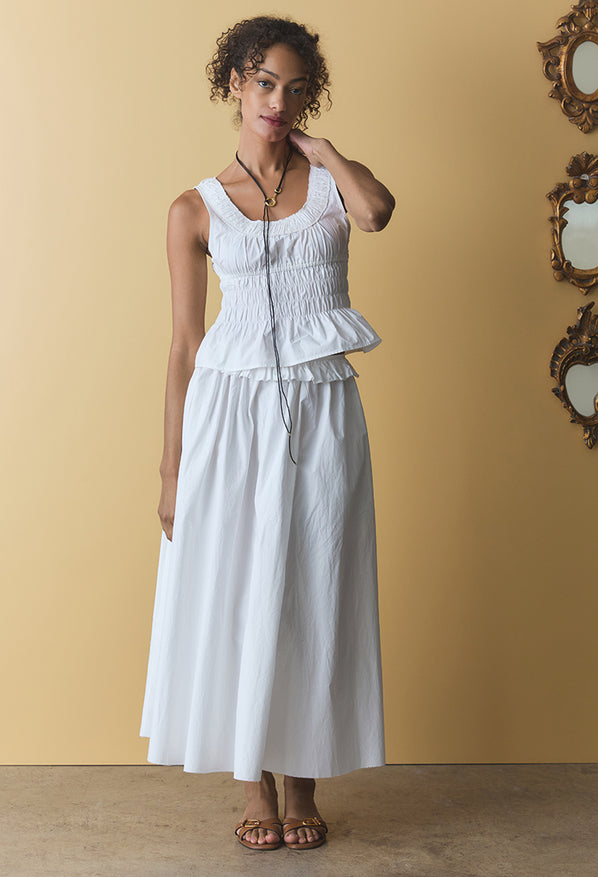 Woman wearing a white sleeveless top with smocking on the waist and long white skirt with ruffles along the waistline against a beige wall.