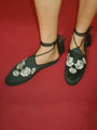 Cropped shin down of woman wearing black slip-on shoes with white floral embroidered in the top and twist-cord ties with tassels wrapped around the ankle on a red background. 