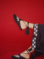 Cropped shin down of woman wearing black slip-on shoes with white floral embroidered in the top and twist-cord ties with tassels wrapped around the ankle on a red background. 