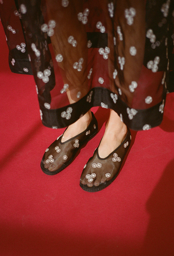 Cropped knee down of woman wearing a sheer black dress with mini white floral embroidery detail and matching sheer slip on black flats on a red background. 