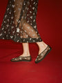 Cropped thigh down of woman wearing a sheer black dress with mini white floral embroidery detail and matching sheer slip on black flats on a red background. 