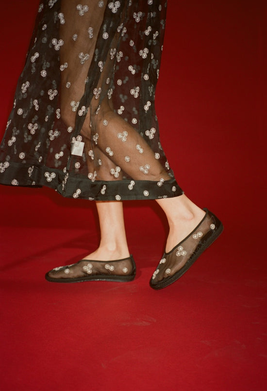 Cropped thigh down of woman wearing a sheer black dress with mini white floral embroidery detail and matching sheer slip on black flats on a red background. 