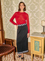 Woman wearing a red sheer knit pullover sweater and black ankle length skirt with small embroidered flowers on the waist and hem standing in a room with patterned wallpaper and furniture.