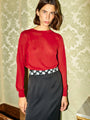 Image cropped to the thigh of a woman wearing a red sheer knit pullover sweater and black ankle length skirt with small embroidered flowers on the waist standing in a room with patterned wallpaper.