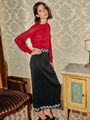 Woman wearing a red sheer knit pullover sweater and black ankle length skirt with small embroidered flowers on the waist and hem standing in a room with patterned wallpaper and furniture.
