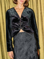 Cropped nose to thigh of woman wearing a black velvet long sleeve blazer top with black floral skirt against a gold curtain background