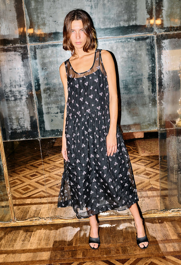 Woman wearing a long black sleeveless sheer dress with small white floral embroidery and drop waist standing in a room with reflective walls and wooden floor.