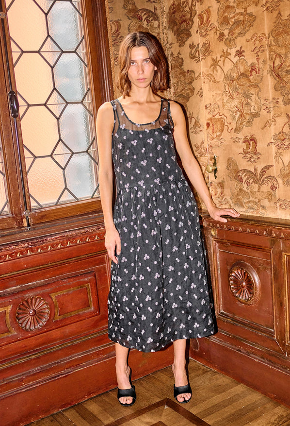 Woman wearing a long black sleeveless sheer dress with small white floral embroidery and drop waist standing in a room with wooden paneling, floral patterned wall and stained glass window.