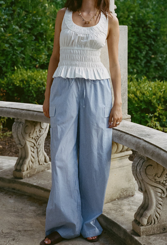 Cropped neck down of woman wearing a white thick strapped tank top with a shirred scoop neckline and tiered rows of smocking around the waist with blue pin-striped pants standing by a stone railing outdoors.