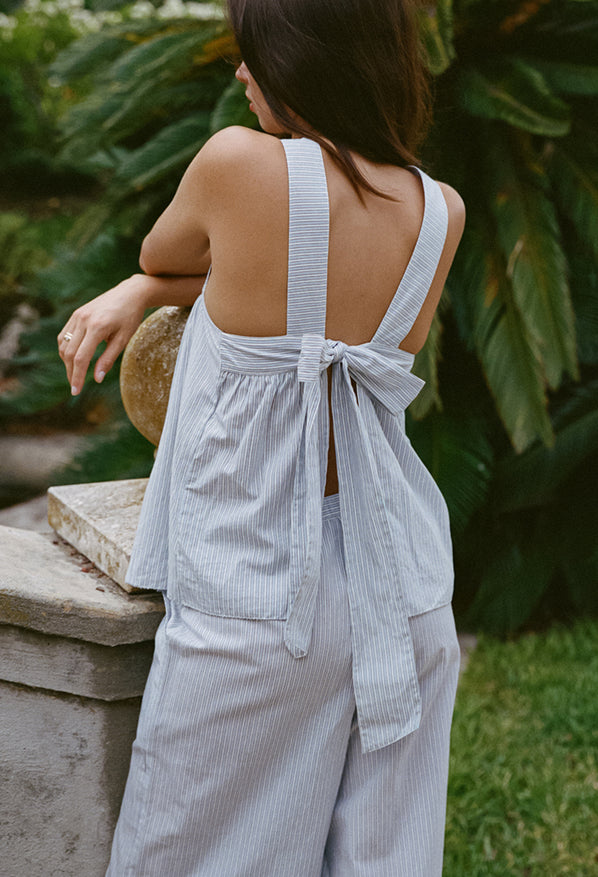 Cropped knees up of woman in light blue pin-striped sleeveless top with tie back and matching wide-leg pants leaning on a stone railing outdoors.