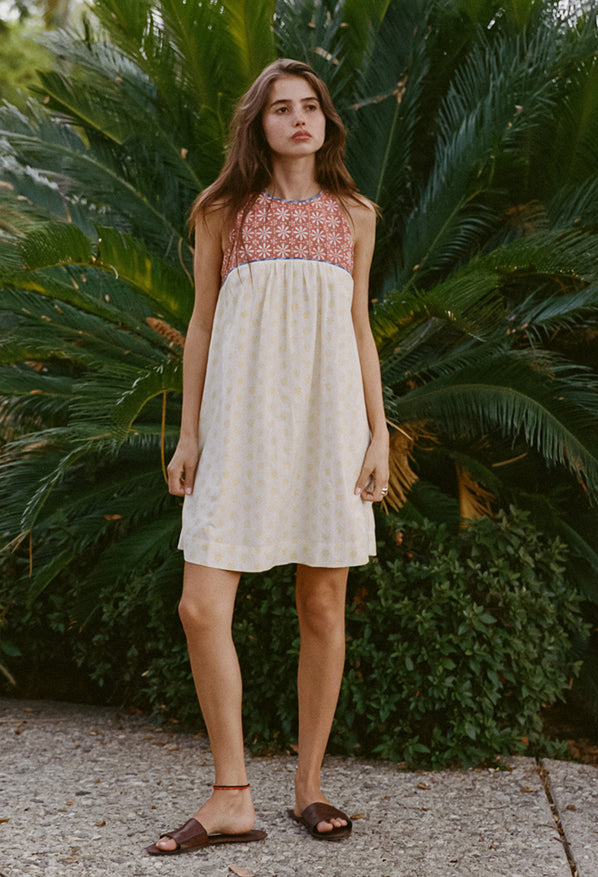 Woman wearing a mini length tank style shift dress with blood orange and white floral top and white skirt with yellow sun pattern standing in front of green foliage