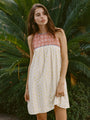 Image cropped knees up of woman wearing a mini length tank style shift dress with blood orange and white floral top and white skirt with yellow sun pattern standing in front of green foliage