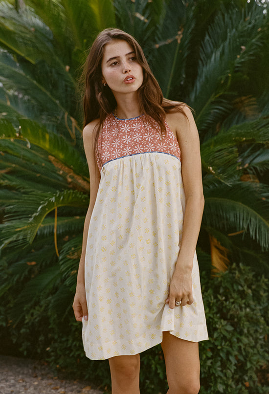 Image cropped knees up of woman wearing a mini length tank style shift dress with blood orange and white floral top and white skirt with yellow sun pattern standing in front of green foliage