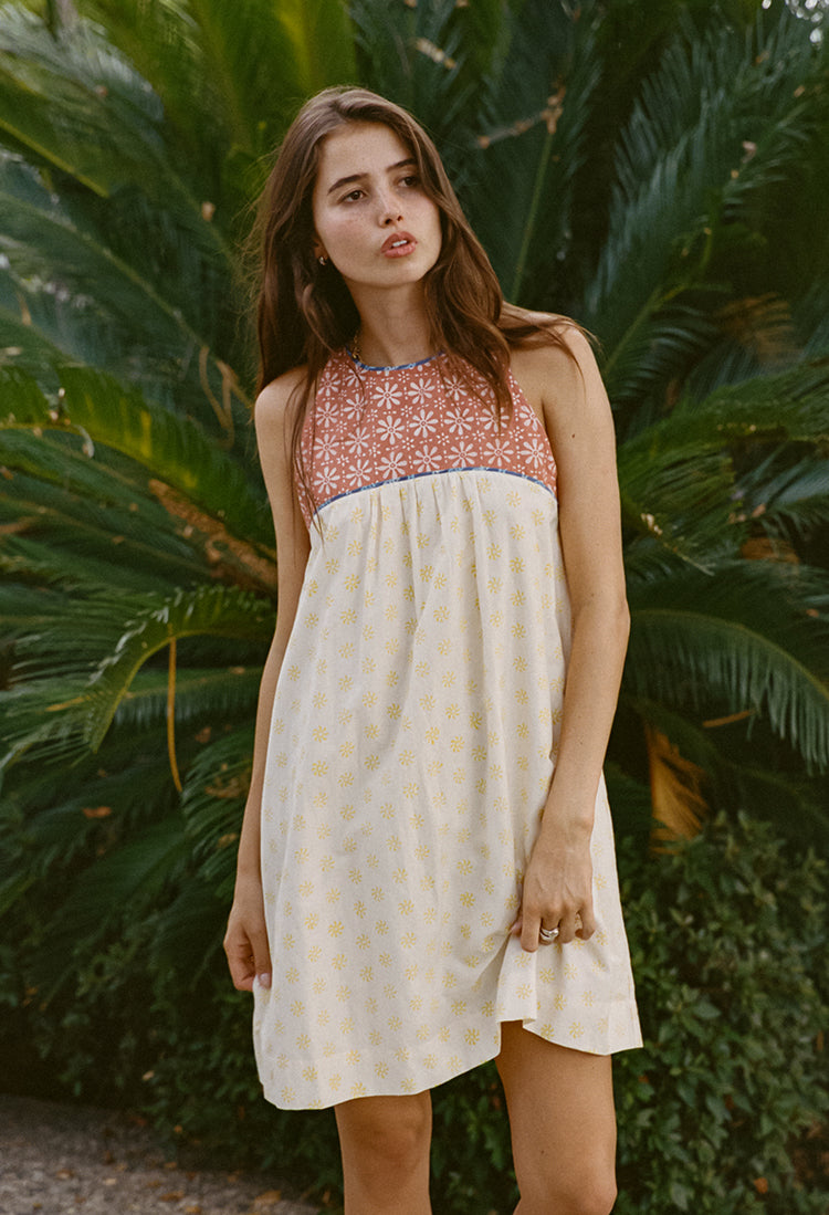 Image cropped knees up of woman wearing a mini length tank style shift dress with blood orange and white floral top and white skirt with yellow sun pattern standing in front of green foliage