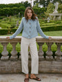 Woman wearing a light blue knitted button down cardigan with chunky braided trim details on the neckline and sleeves and ivory pants with a garden in the background.