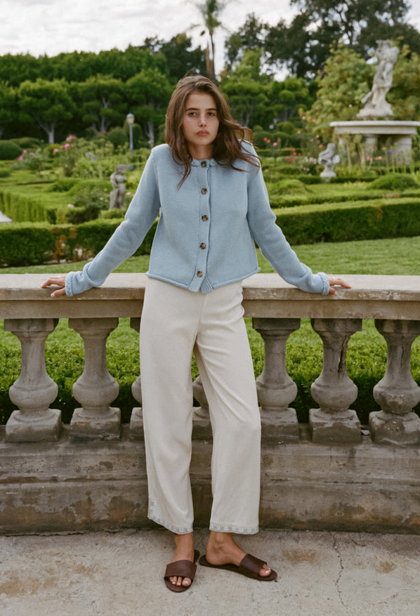 Woman wearing a light blue knitted button down cardigan with chunky braided trim details on the neckline and sleeves and ivory pants with a garden in the background.