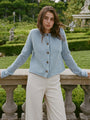 Cropped knees up of woman wearing a light blue knitted button down cardigan with chunky braided trim details on the neckline and sleeves and ivory pants with a garden in the background.