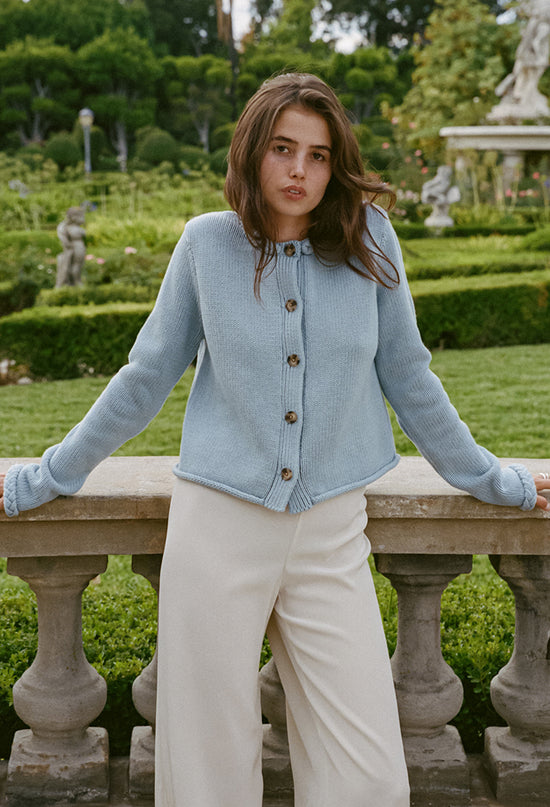 Cropped knees up of woman wearing a light blue knitted button down cardigan with chunky braided trim details on the neckline and sleeves and ivory pants with a garden in the background.