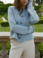 Cropped eyes to thigh of woman wearing a light blue knitted button down cardigan with chunky braided trim details on the neckline and sleeves and ivory pants with a garden in the background.