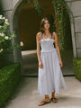 Woman in a light blue pin-striped midi length dress with spaghetti straps, smocked bodice and ruffled neckline and waistline standing in front of an archway with greenery
