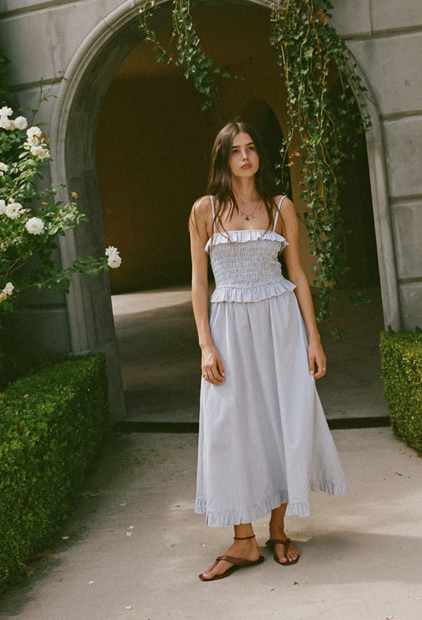 Woman in a light blue pin-striped midi length dress with spaghetti straps, smocked bodice and ruffled neckline and waistline standing in front of an archway with greenery