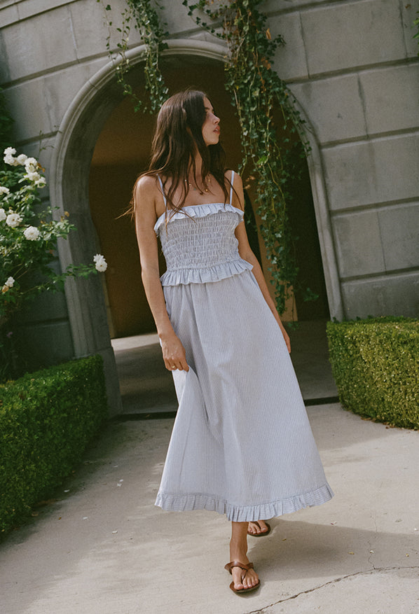 Woman in a light blue pin-striped midi length dress with spaghetti straps, smocked bodice and ruffled neckline and waistline walking in front of a stone building with greenery.