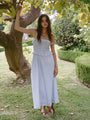 Woman in a light blue pin-striped midi length dress with spaghetti straps, smocked bodice and ruffled neckline and waistline standing under a tree in a garden