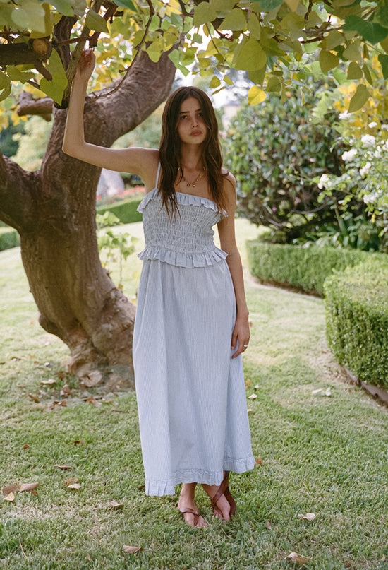Woman in a light blue pin-striped midi length dress with spaghetti straps, smocked bodice and ruffled neckline and waistline standing under a tree in a garden