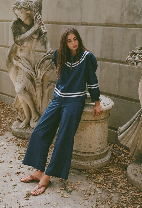 Woman in a navy blue outfit leaning against a stone pedestal next to stone statues and a column.