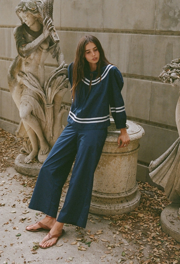 Woman in a navy blue outfit leaning against a stone pedestal next to stone statues and a column.