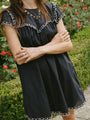 Cropped chin to knee of woman wearing a black mini dress with cap sleeves and white floral embroidery across the chest, sleeve and hemline standing in an outdoor setting.