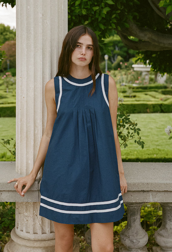 Cropped knee up of woman in a sleeveless blue short dress with white neck, sleeve, and hem detailing standing in a garden setting