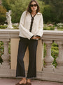Woman in ivory knit jacket with black trim detailing along the sleeves, hem, and center seam and black pants standing in a garden setting.