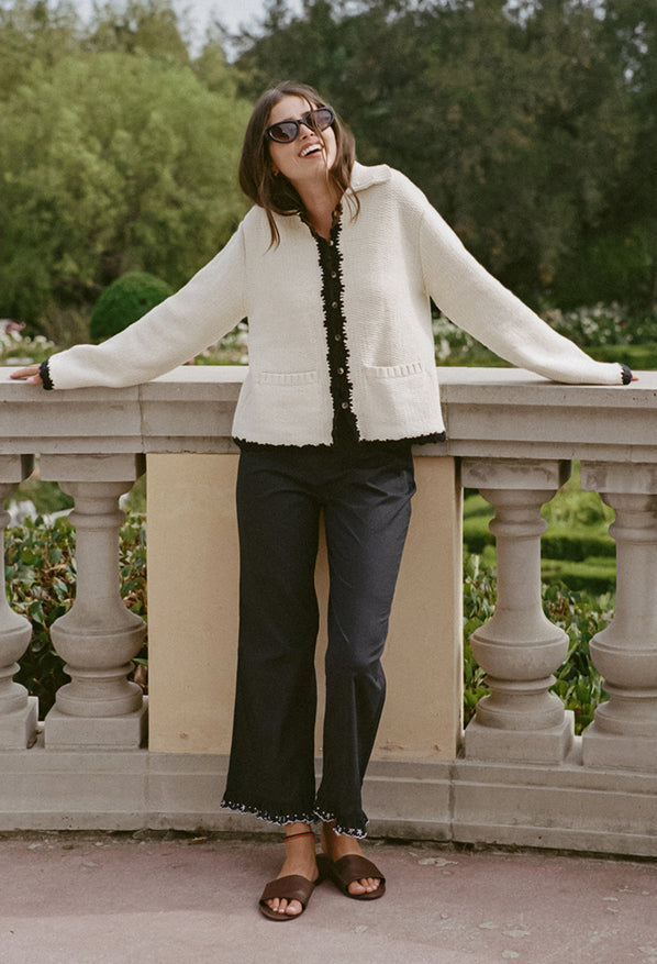 Woman in ivory knit jacket with black trim detailing along the sleeves, hem, and center seam and black pants standing in a garden setting.