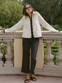 Woman in ivory knit jacket with black trim detailing along the sleeves, hem, and center seam and black pants standing in a garden setting.