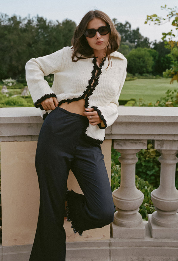 Woman in ivory knit jacket with black trim detailing along the sleeves, hem, and center seam and black pants standing in a garden setting against a stone balcony.