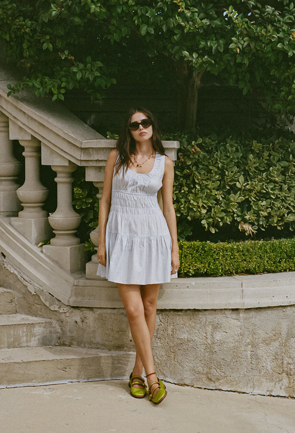 Woman in a light blue pin-striped tank style mini dress with a smocked bodice standing outdoors with greenery in the background