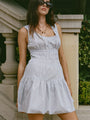 Image cropped head to knee of woman in a light blue pin-striped tank style mini dress with a smocked bodice standing outdoors with greenery in the background
