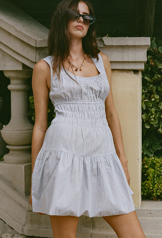 Image cropped head to knee of woman in a light blue pin-striped tank style mini dress with a smocked bodice standing outdoors with greenery in the background
