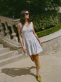 Woman in a light blue pin-striped tank style mini dress with a smocked bodice walking outdoors with stone steps and greenery in the background
