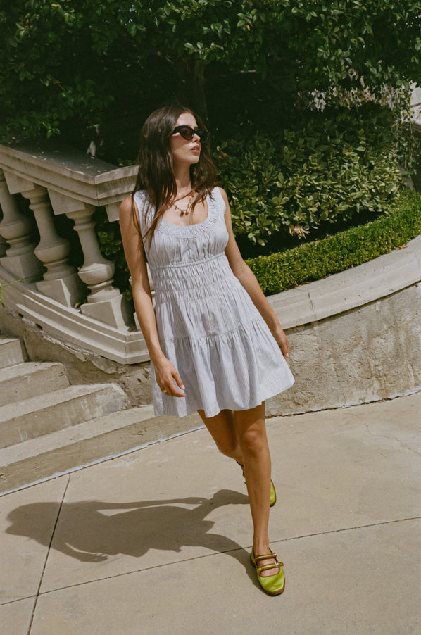 Woman in a light blue pin-striped tank style mini dress with a smocked bodice walking outdoors with stone steps and greenery in the background