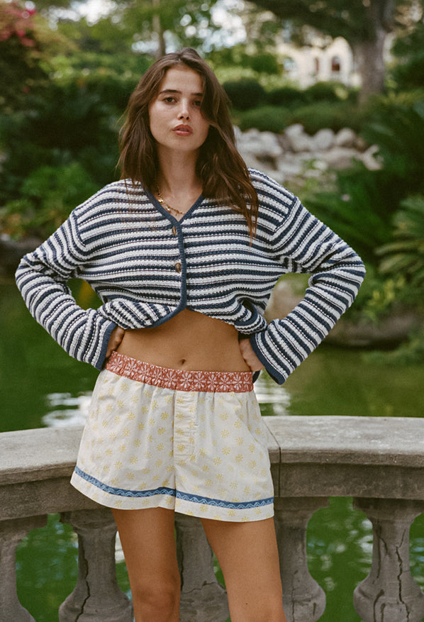Cropped knees up of woman wearing a striped white and navy cardigan and shorts with blood orange floral top waistband and white legs with yellow sun pattern and blue hem detail standing outdoors.