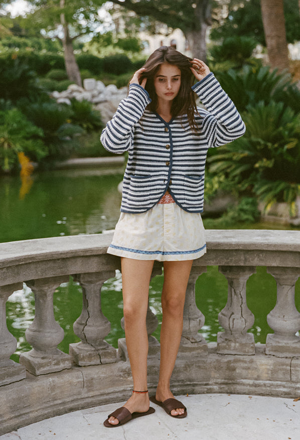 Woman wearing a striped white and navy cardigan and shorts with blood orange floral top waistband and white legs with yellow sun pattern and blue hem detail standing against a stone railing outdoors.