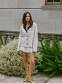 Woman wearing an off white jacket with black embroidery detailing, black collar and dark horn buttons and silky white shorts on standing outdoors in front of a building with plants