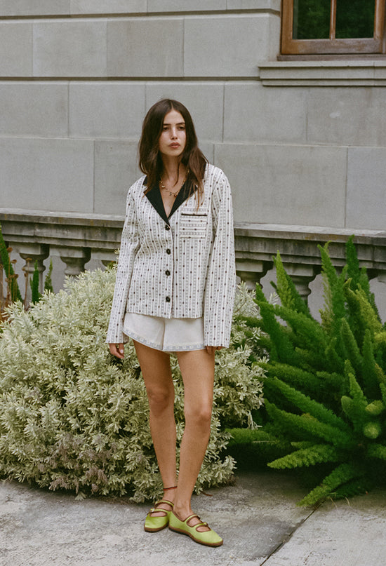 Woman wearing an off white jacket with black embroidery detailing, black collar and dark horn buttons and silky white shorts on standing outdoors in front of a building with plants