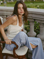Women wearing a cream knitted button up tank top with black hem details paired with light blue pin-striped pants sitting on a chair outdoors with a blurred background