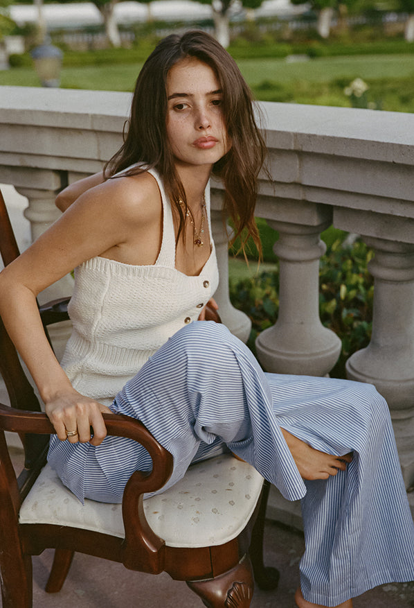 Women wearing a cream knitted button up tank top with black hem details paired with light blue pin-striped pants sitting on a chair outdoors with a blurred background