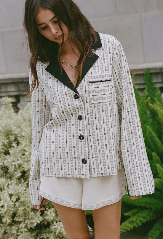 Cropped knees up of woman wearing an off white jacket with black embroidery detailing, black collar and dark horn buttons and silky white shorts on standing outdoors in front of a building with plants