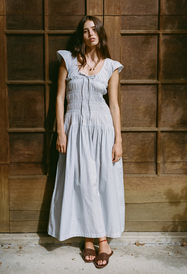Woman wearing a light blue pinstriped short-sleeved midi dress with a scoop neckline with a tie and smocking around the bust to the waist standing against a wooden wall.