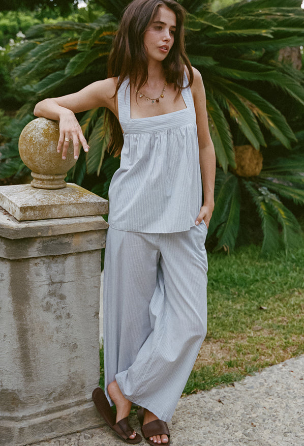 Woman in light blue pin-striped sleeveless top and matching wide-leg pants leaning on a stone railing outdoors.